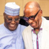 Two friends smiling and posing together, one wearing a patterned hat and striped shirt, the other in a light blazer and glasses.