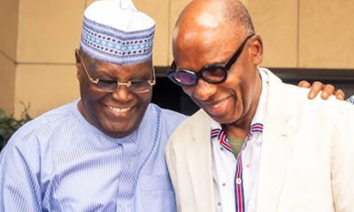 Two friends smiling and posing together, one wearing a patterned hat and striped shirt, the other in a light blazer and glasses.