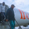 Two men stepping off a white aircraft via portable stairs on a tarmac, with orange lettering along the fuselage.