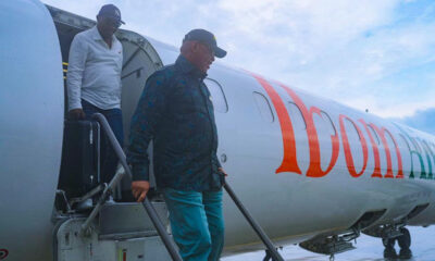 Two men stepping off a white aircraft via portable stairs on a tarmac, with orange lettering along the fuselage.