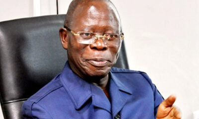 Older man with glasses in a blue uniform jacket sits in a black office chair, speaking and gesturing with his right hand.