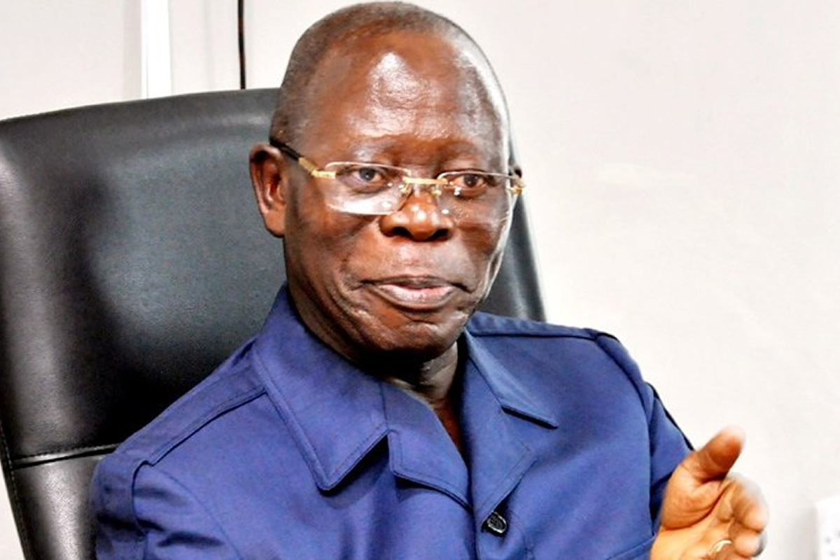 Older man with glasses in a blue uniform jacket sits in a black office chair, speaking and gesturing with his right hand.