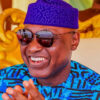 Smiling man wearing a blue patterned shirt and a purple traditional hat with sunglasses in a bright, decorative setting.