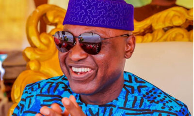Smiling man wearing a blue patterned shirt and a purple traditional hat with sunglasses in a bright, decorative setting.