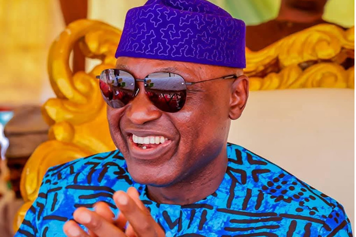 Smiling man wearing a blue patterned shirt and a purple traditional hat with sunglasses in a bright, decorative setting.