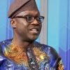 Man wearing a colorful traditional African print shirt and brown cap, speaking or mid-conversation, with glasses on and a blue geometric background.