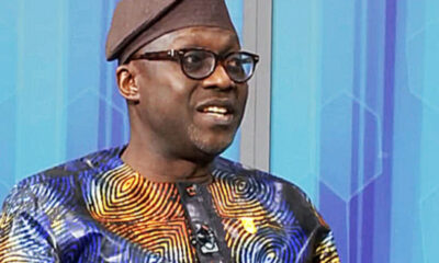 Man wearing a colorful traditional African print shirt and brown cap, speaking or mid-conversation, with glasses on and a blue geometric background.
