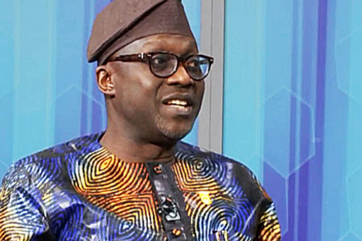 Man wearing a colorful traditional African print shirt and brown cap, speaking or mid-conversation, with glasses on and a blue geometric background.