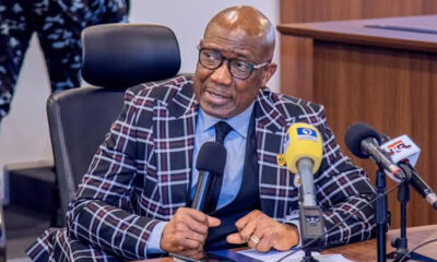 Man in a plaid suit sits at a table with microphones during a press conference.