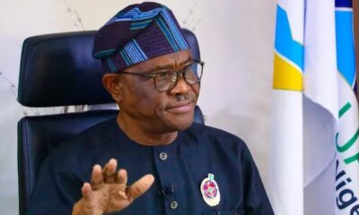 Nigerian man in dark traditional attire and patterned cap sits in an executive chair, gesturing with his hand during a formal event, flags in the background.