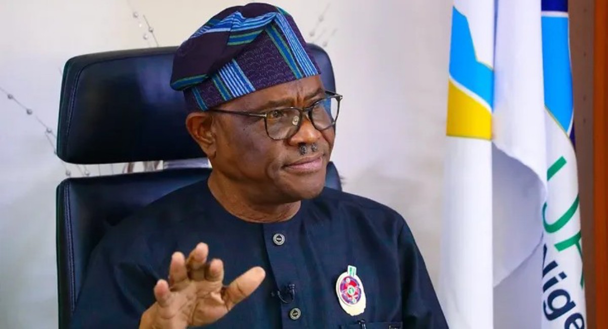 Nigerian man in dark traditional attire and patterned cap sits in an executive chair, gesturing with his hand during a formal event, flags in the background.