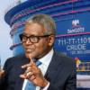 Businessman wearing glasses in a dark suit and blue tie gestures while speaking, with a large Dangote crude oil storage tank in the background.