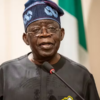 Nigerian man in traditional dark agbada and patterned cap speaks at a microphone, with a flag in the background.