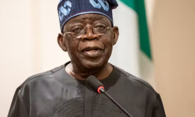Nigerian man in traditional dark agbada and patterned cap speaks at a microphone, with a flag in the background.