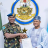 Two men shake hands, holding a signed document, in front of a Nigerian Air Force emblem and flags behind them with a star in the center badge of the force.