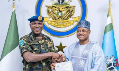 Two men shake hands, holding a signed document, in front of a Nigerian Air Force emblem and flags behind them with a star in the center badge of the force.