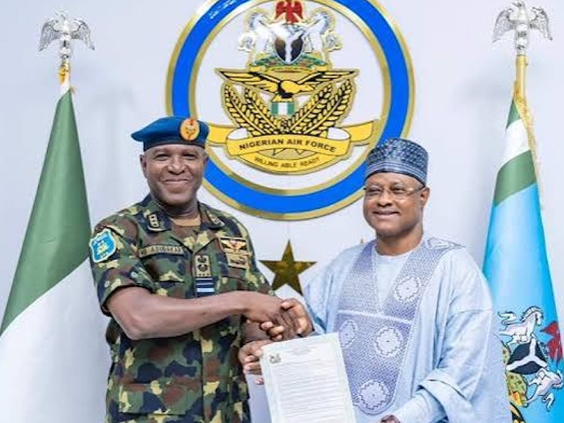 Two men shake hands, holding a signed document, in front of a Nigerian Air Force emblem and flags behind them with a star in the center badge of the force.