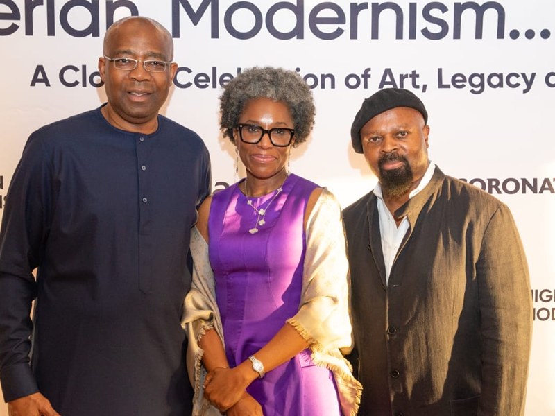 Three people pose for a photo at an art celebration event, with a banner behind them that reads 'Modernism... A Celebration of Art, Legacy'.
