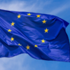 Blue European Union flag fluttering in the wind against a clear blue sky, with 12 yellow stars in a circle.