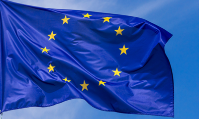 Blue European Union flag fluttering in the wind against a clear blue sky, with 12 yellow stars in a circle.