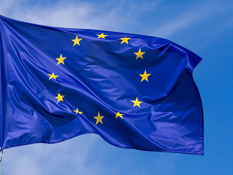 Blue European Union flag fluttering in the wind against a clear blue sky, with 12 yellow stars in a circle.