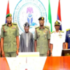 Group of military officers and a civilian in traditional attire standing behind a desk during a formal ceremony, with a large government seal and national flags in the background.