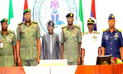 Group of military officers and a civilian in traditional attire standing behind a desk during a formal ceremony, with a large government seal and national flags in the background.