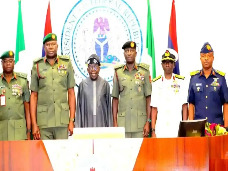 Group of military officers and a civilian in traditional attire standing behind a desk during a formal ceremony, with a large government seal and national flags in the background.