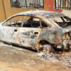 Charred, destroyed sedan with burnt exterior and melted wheels, parked beside a wall after a fire.