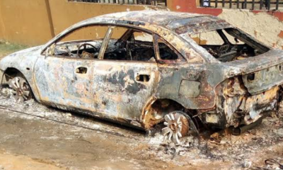 Charred, destroyed sedan with burnt exterior and melted wheels, parked beside a wall after a fire.