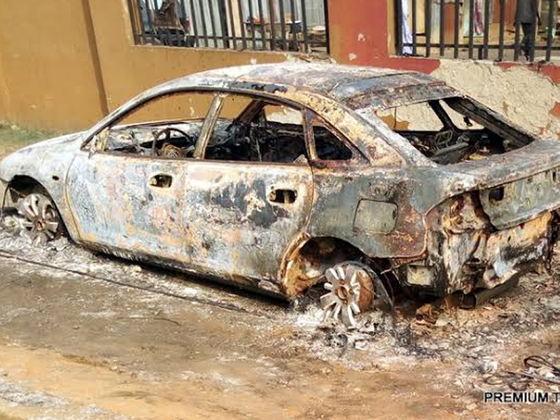 Charred, destroyed sedan with burnt exterior and melted wheels, parked beside a wall after a fire.