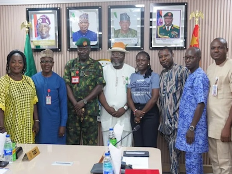 Group of ten adults in an office posing for a formal photo, including a military officer and officials, with framed portraits on the wall behind them.