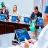 Delegates in a formal meeting sit at a long table with laptops and flags.