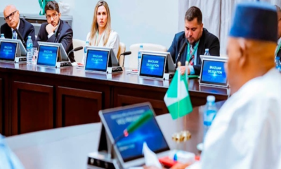 Delegates in a formal meeting sit at a long table with laptops and flags.