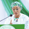 Woman wearing a green patterned head wrap and glasses speaks at a green podium labeled 'First Lady' with a microphone.