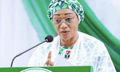 Woman wearing a green patterned head wrap and glasses speaks at a green podium labeled 'First Lady' with a microphone.