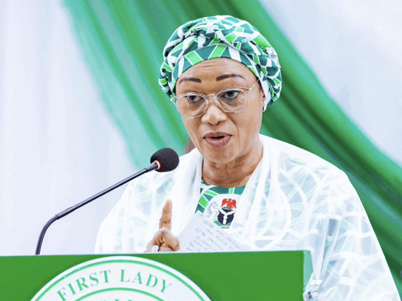 Woman wearing a green patterned head wrap and glasses speaks at a green podium labeled 'First Lady' with a microphone.