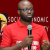 Man with glasses and a beard in a red polo shirt speaking into a handheld microphone on a red backdrop with a logo.