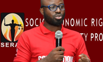 Man with glasses and a beard in a red polo shirt speaking into a handheld microphone on a red backdrop with a logo.