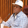 Man in a blue-checkered shirt and white cap speaking into a microphone at a meeting table.