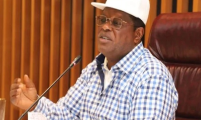 Man in a blue-checkered shirt and white cap speaking into a microphone at a meeting table.