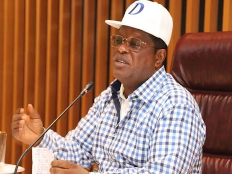 Man in a blue-checkered shirt and white cap speaking into a microphone at a meeting table.