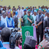 Politician in green attire speaks at a podium with Nigerian emblem, flanked by supporters and security while reporters film the event.