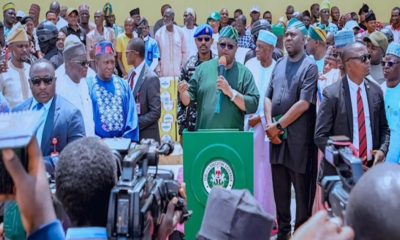 Politician in green attire speaks at a podium with Nigerian emblem, flanked by supporters and security while reporters film the event.