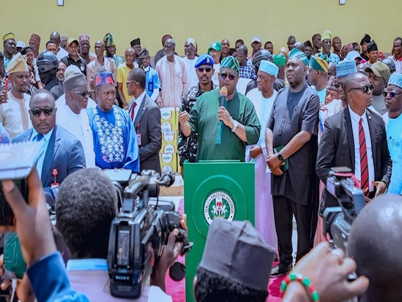 Politician in green attire speaks at a podium with Nigerian emblem, flanked by supporters and security while reporters film the event.