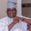 Smiling man in traditional white-and-silver agbada and cap, seated on a sofa indoors.