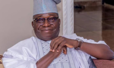 Smiling man in traditional white-and-silver agbada and cap, seated on a sofa indoors.