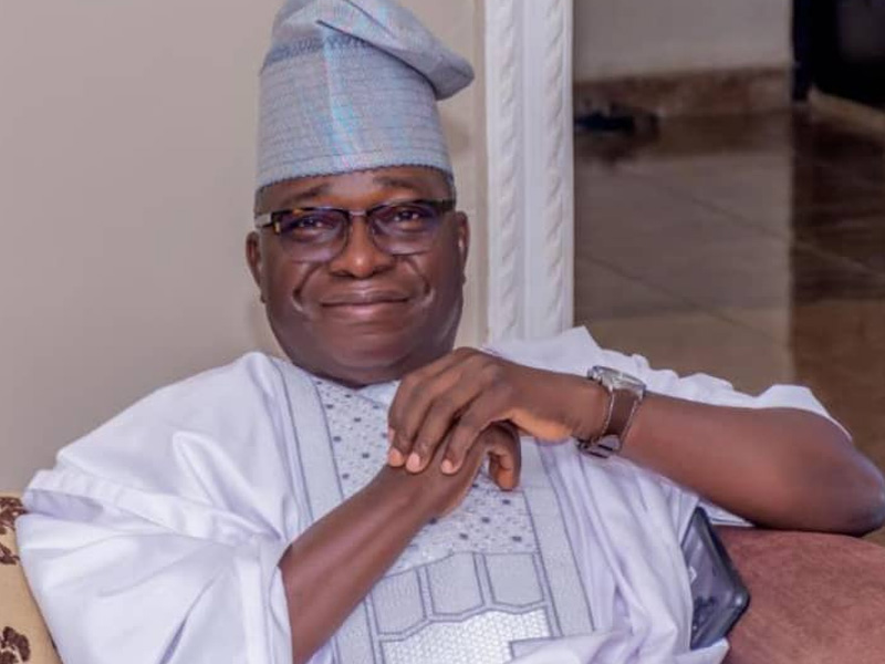 Smiling man in traditional white-and-silver agbada and cap, seated on a sofa indoors.