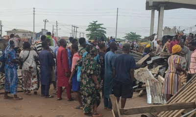 Crowd of people in colorful clothing gathered around rubble and debris on a dusty street after a collapse, with a raised structure visible in the background.