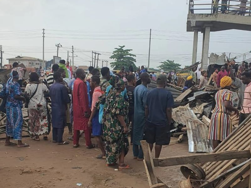 Crowd of people in colorful clothing gathered around rubble and debris on a dusty street after a collapse, with a raised structure visible in the background.